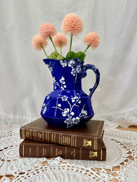 Blue floral pitcher with pink flowers on stacked books against a white lace tablecloth.