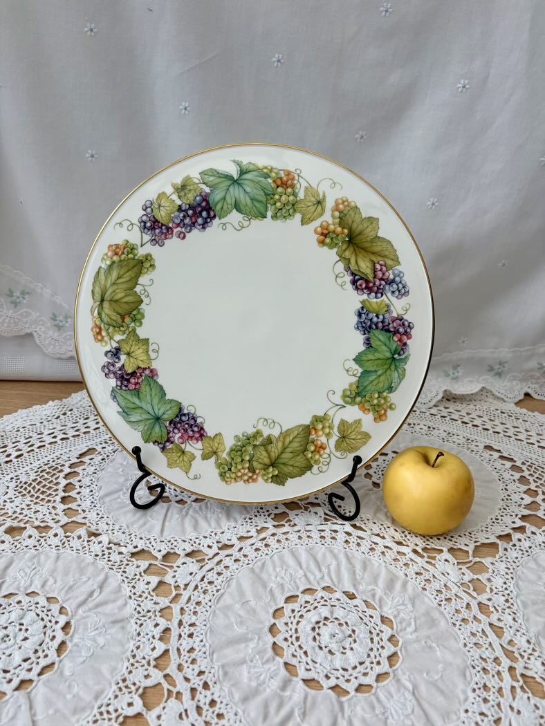 Decorative plate with grapevine design on a stand next to a gold apple on a lace tablecloth.