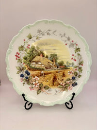 Decorative plate with a scenic design of a house and nature on a stand against a plain background.