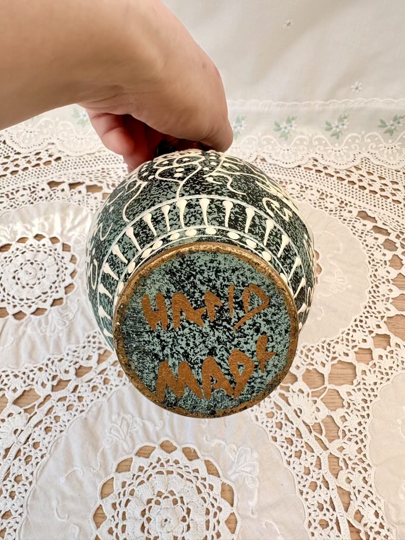 Hand holding a textured ceramic jar with 'Hand made' inscription on a lace tablecloth.