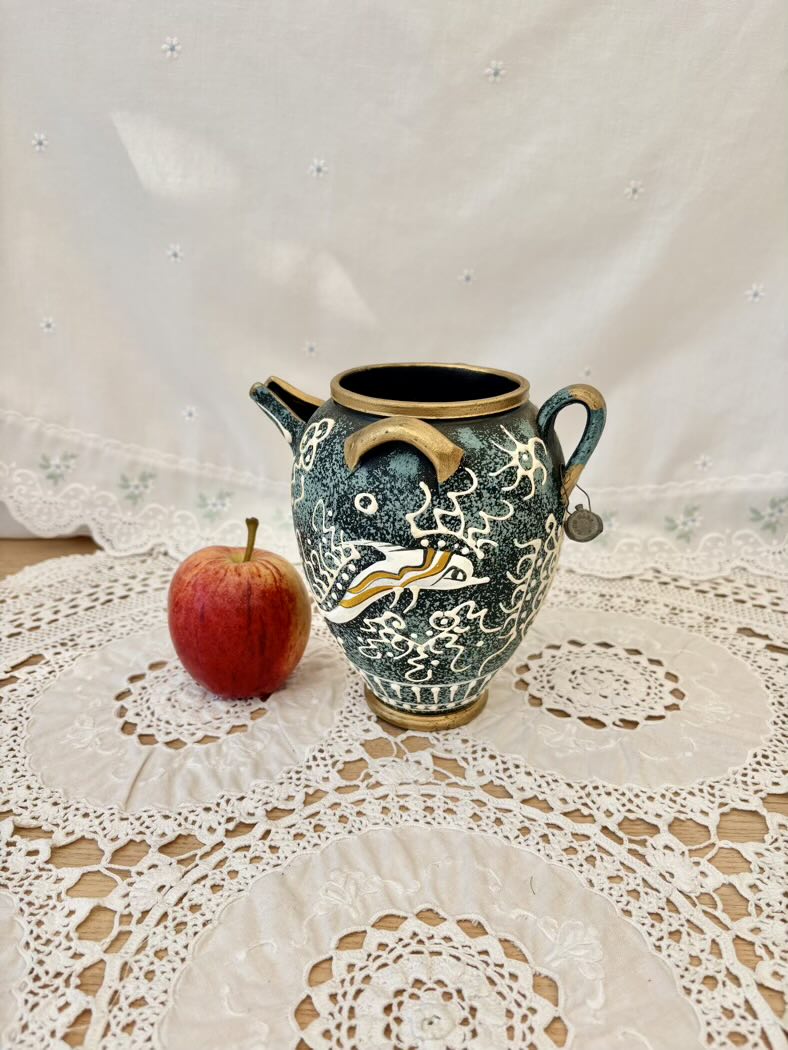 Decorative ceramic pitcher with intricate designs on a lace tablecloth with an apple beside it.
