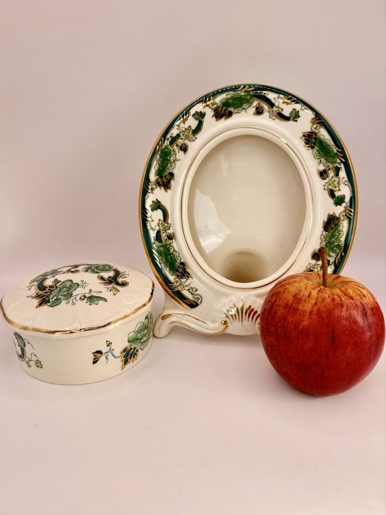 Decorative ceramic bowl with lid and floral patterns next to an apple on a plain background