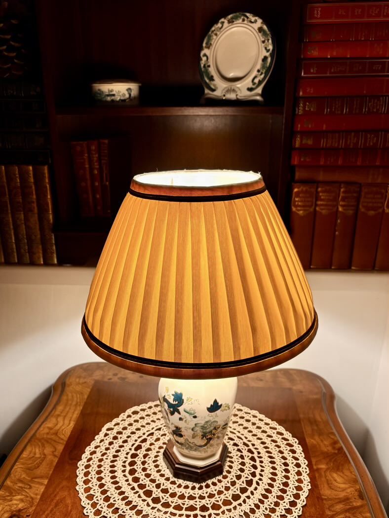Table lamp with pleated yellow shade on a wooden table, with books and decorative items in the background.