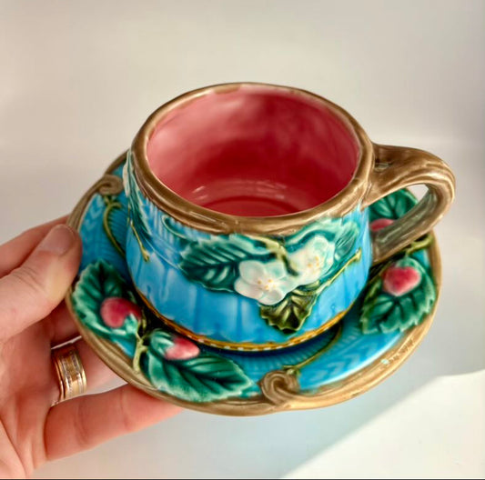 Decorative ceramic cup and saucer with floral design held by a hand against a neutral background