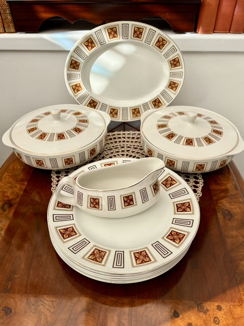 Vintage bone china dinner service set displayed on a wooden sideboard, featuring a stack of dinner plates with a gravy boat on top, two lidded tureens, and an oval platter, all with matching geometric dark red, gold and black diamond border pattern