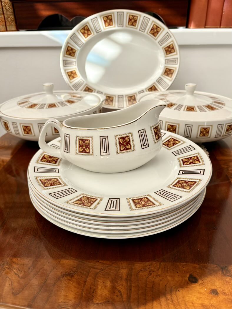 Close-up of a vintage bone china dinner service set on a walnut sideboard, showing a stack of dinner plates with a gravy boat resting on top, two lidded tureens and an oval platter in the background, all featuring a bold geometric border with dark red floral diamond motifs and gold and black rectangular frames