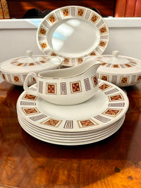 Close-up of a vintage bone china dinner service set on a walnut sideboard, showing a stack of dinner plates with a gravy boat resting on top, two lidded tureens and an oval platter in the background, all featuring a bold geometric border with dark red floral diamond motifs and gold and black rectangular frames