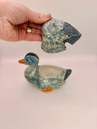 Ceramic duck-shaped object with a hand holding a crystal above it on a light background