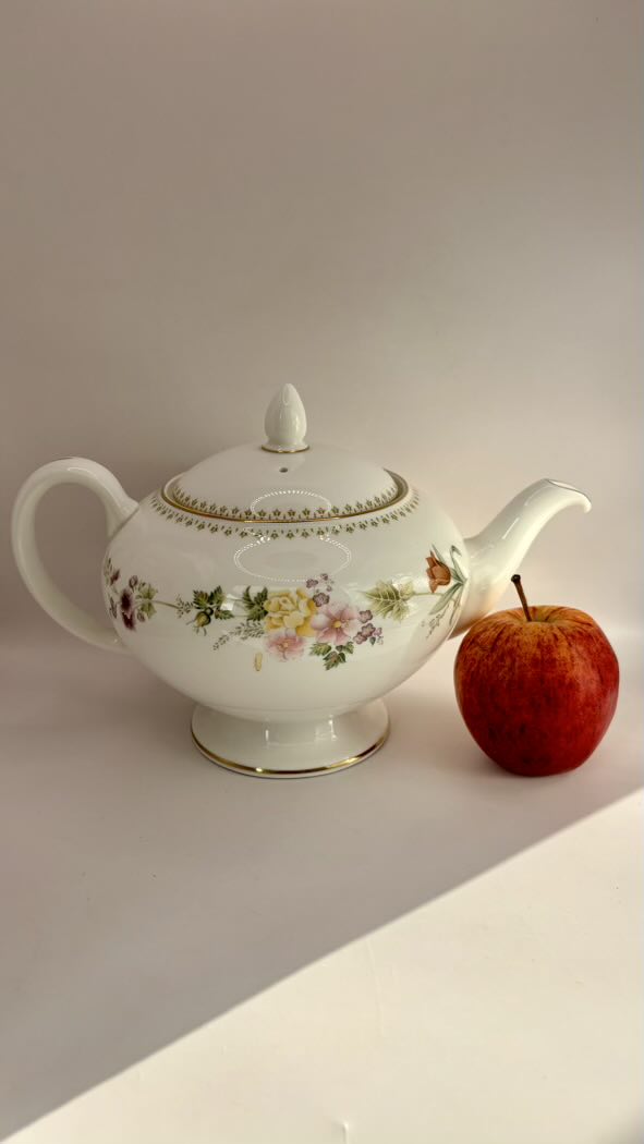 Decorative teapot with floral patterns next to an apple on a light surface.