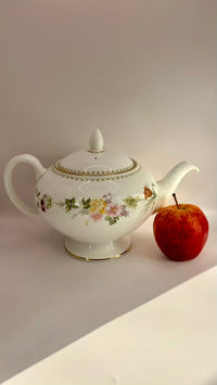 Decorative teapot with floral patterns next to an apple on a light surface.