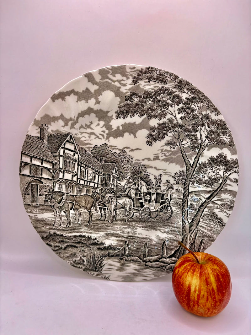 Decorative plate with black and white landscape design featuring a village scene, next to an apple on a white background.