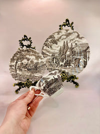 Decorative teacup with scenic design and ornate handles held by a hand against a light background