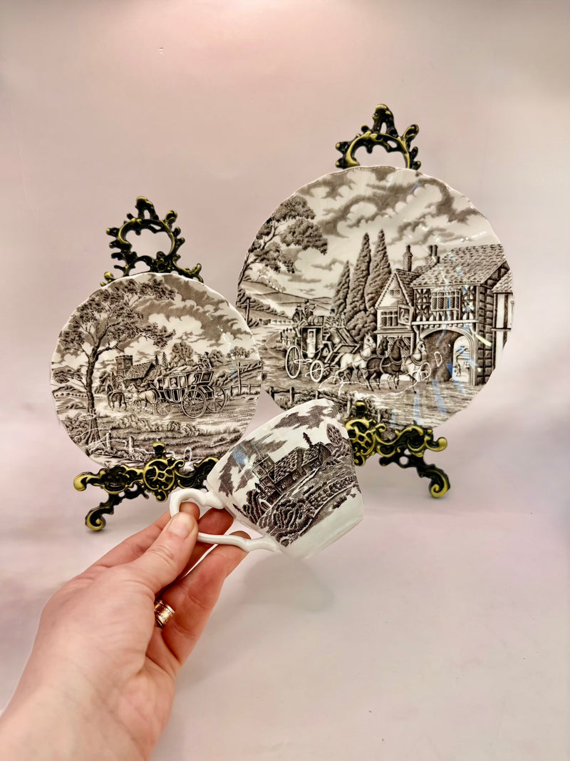 Decorative teacup with scenic design and ornate handles held by a hand against a light background