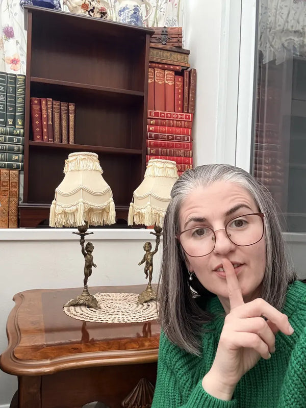 Woman in a green sweater with a finger over her lips in front of a bookshelf with decorative items.