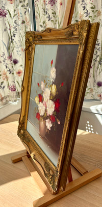 Decorative gold-framed painting of flowers on a wooden easel with floral curtains in the background.