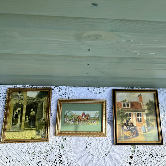 Three framed paintings on a lace tablecloth with a wooden surface above.