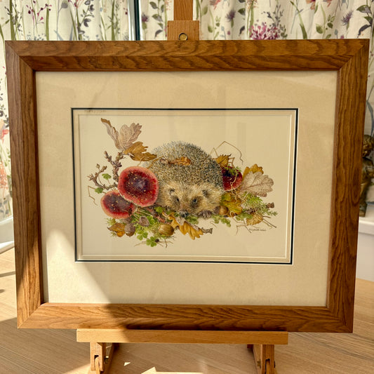 Framed artwork of a hedgehog among flowers and leaves on a wooden easel.