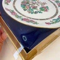 Decorative plate with floral design on a blue cushion inside a cardboard box.