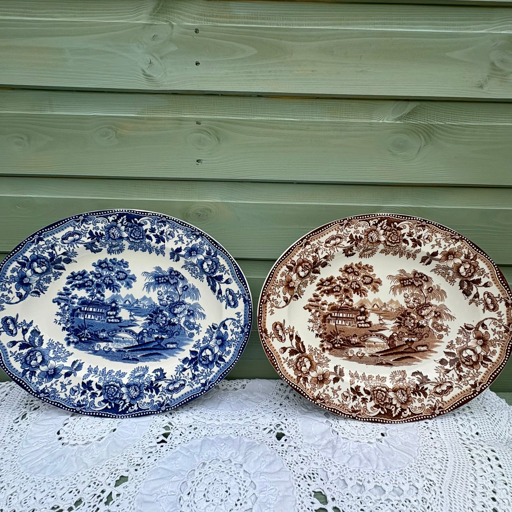 Two decorative plates with blue and brown patterns on a lace tablecloth against a wooden background.