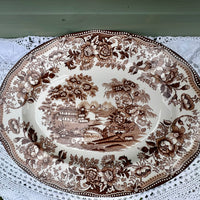 Decorative plate with brown floral and landscape design on a white lace tablecloth.