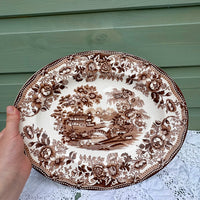 Decorative plate with brown floral and landscape design held by a hand against a green wooden background.