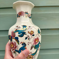 Decorative vase with floral and bird patterns held by a hand against a neutral background