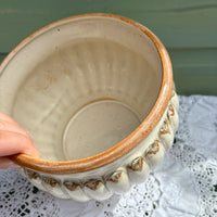 Ceramic pot with decorative edge held by a hand on a lace tablecloth.