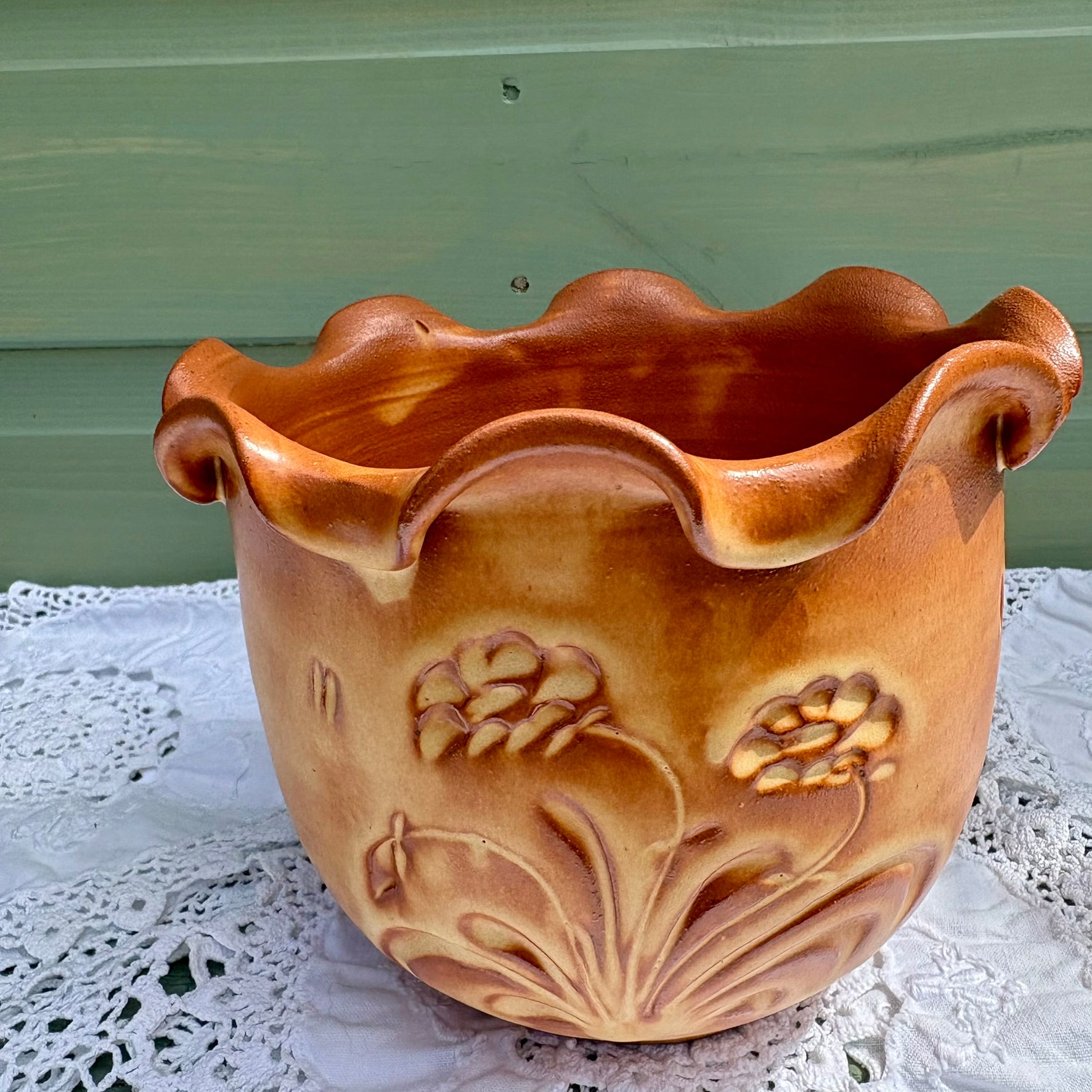Ceramic planter with floral designs on a lace tablecloth against a green wall.