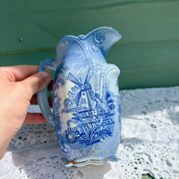 Blue and white ceramic pitcher with a windmill design held by a hand on a lace tablecloth.