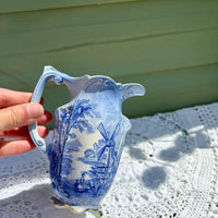 Blue and white ceramic pitcher with windmill design held by a hand on a lace tablecloth.