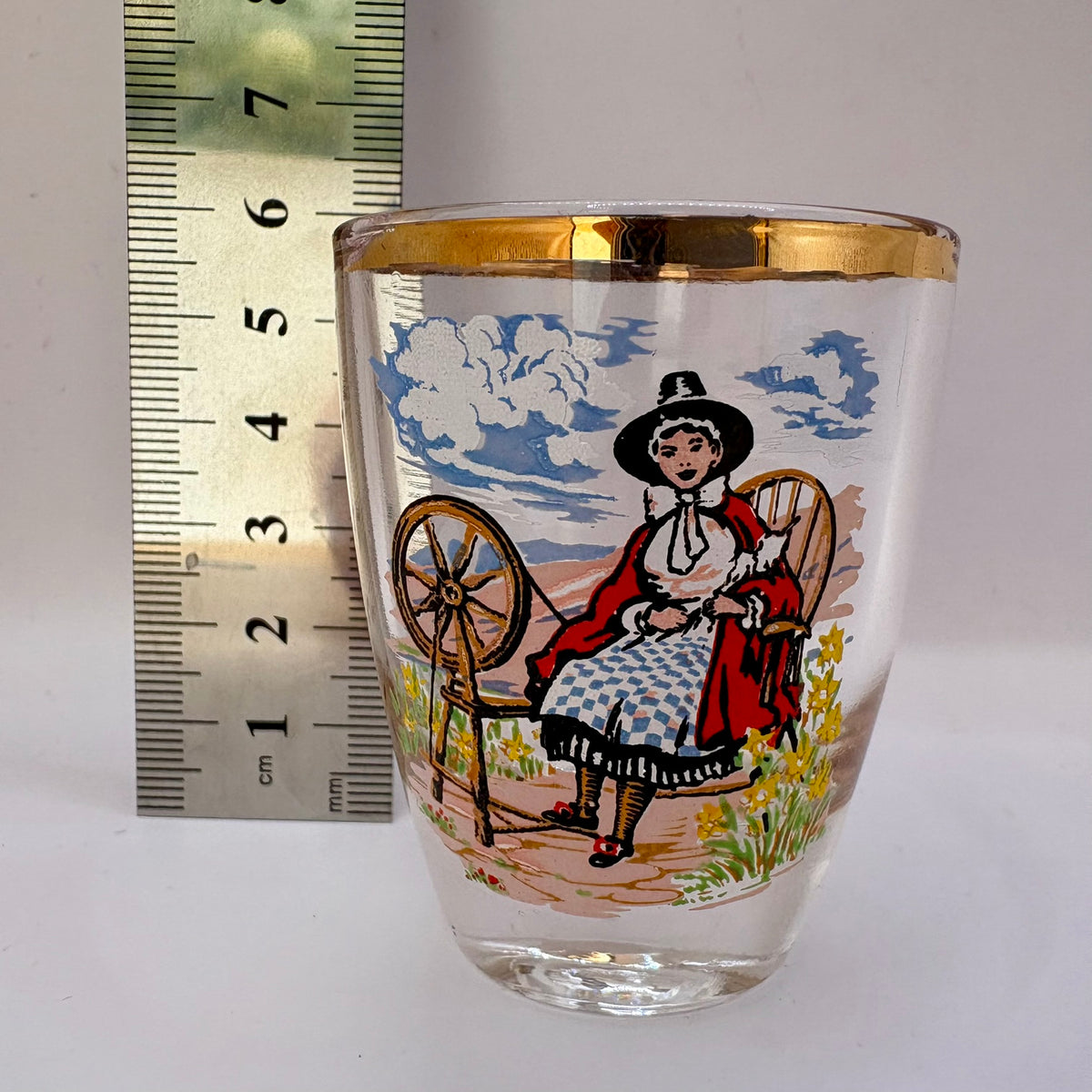 Decorative glass with a printed design of a woman spinning, next to a ruler for scale.