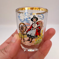 Glass shot glass with a decorative design of a woman with a spinning wheel held by a hand.