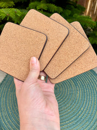 Cork coasters held by a hand with a ring, on a textured surface with greenery in the background.