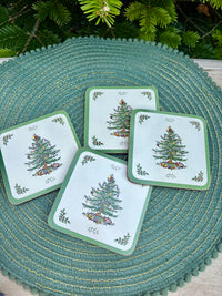 Set of four Christmas-themed coasters on a green woven mat with evergreen branches in the background.