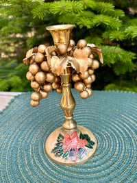Decorative candle holder with gold grapes on a textured surface with greenery in the background