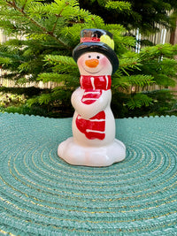 Snowman figurine with a red scarf on a textured surface with greenery in the background