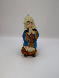 Small figurine of a person in blue clothing with a white beard, standing over a manger with a baby in it, on a white background.