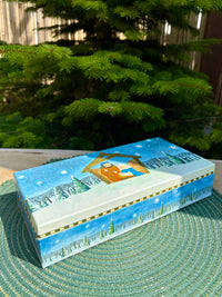 Decorative box with a winter scene painting on a textured surface with greenery in the background