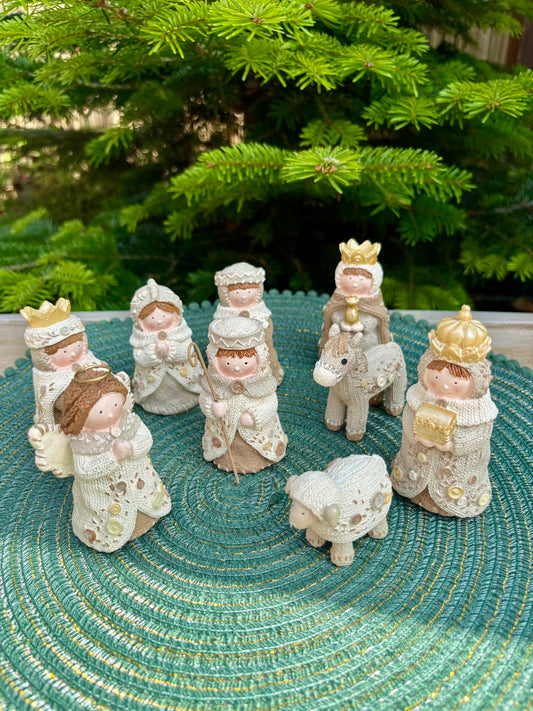 Nativity scene figurines on a textured surface with a green woven mat and blurred Christmas tree in the background.