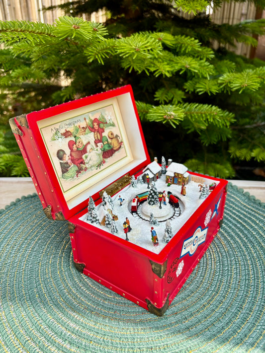 Red toy box with a Christmas scene and animated figures in front of a Christmas tree.