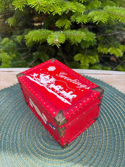 Red decorative box with 'Greetings' text on a green textured surface with a blurred Christmas tree in the background.