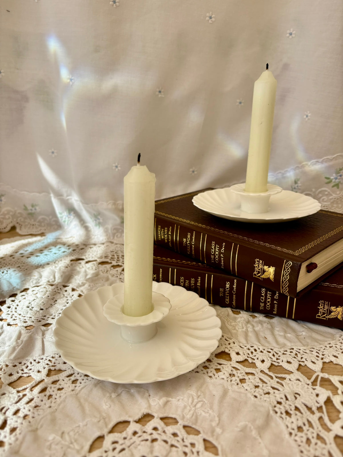 Two white candles in decorative holders on a lace tablecloth with books in the background.