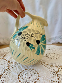 Ceramic pitcher with floral design held by a hand on a lace tablecloth.