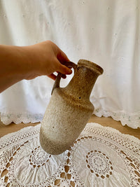 Hand holding a textured ceramic vase on a lace tablecloth with a white curtain background