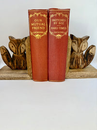 Two red books titled 'Our Mutual Friend' and 'Sketches by Boz' by Charles Dickens, flanked by wooden fox figures on a white background.
