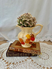 Decorative pitcher with autumn leaves on a stack of books, placed on a lace tablecloth.