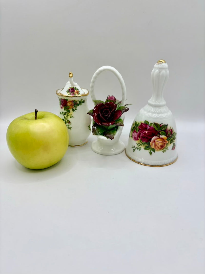 Decorative ceramic items including a bell, basket, and jar with floral designs next to a green apple on a white background.