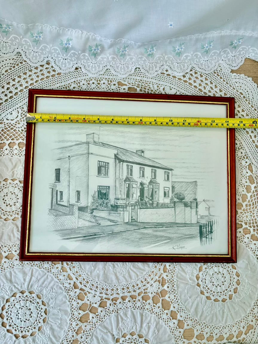 Framed black and white drawing of a house on a lace tablecloth with a measuring tape.
