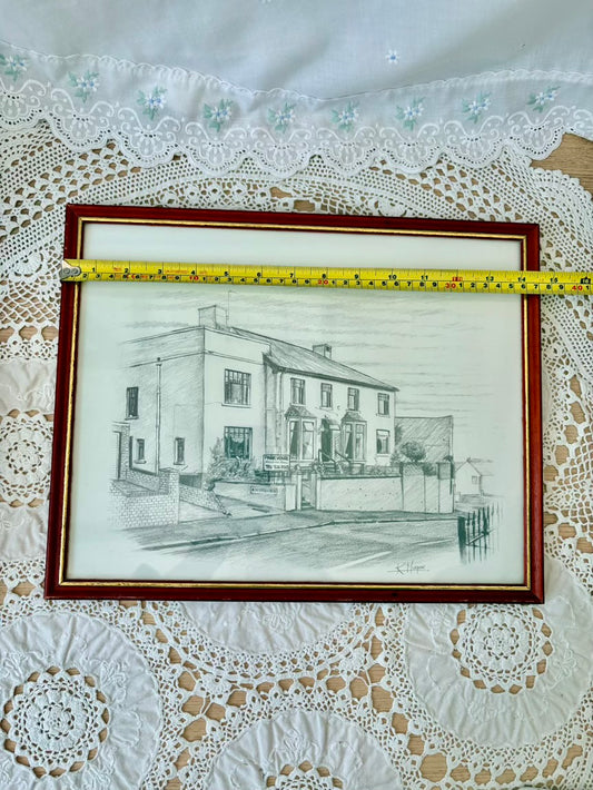 Framed black and white drawing of a house on a lace tablecloth with a measuring tape.