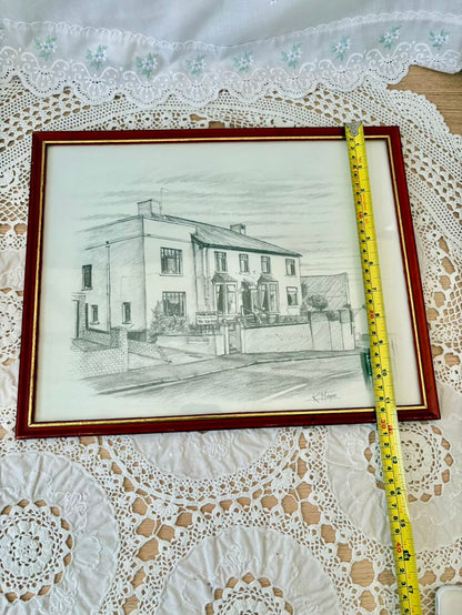 Framed pencil drawing of a house on a lace tablecloth with a measuring tape.
